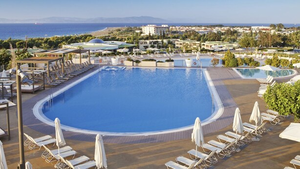 A large pool with sun loungers and umbrellas, surrounded by buildings and a view of the sea. - Kipriotis Panorama and Suites (Kos)