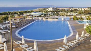 A large pool with sun loungers and umbrellas, surrounded by buildings and a view of the sea.