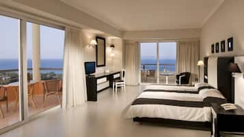 A modern hotel room with a large bed, a desk, a TV, and a balcony with a view of the sea.