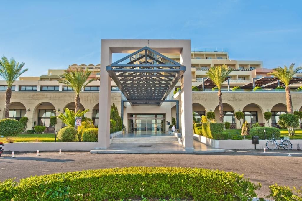A modern hotel entrance with a geometric canopy, arched windows, and palm trees.