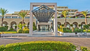 A modern hotel entrance with a geometric canopy, arched windows, and palm trees. - Kipriotis Panorama and Suites (Kos)