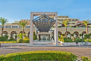 A modern hotel entrance with a geometric canopy, arched windows, and palm trees. - Kipriotis Panorama and Suites (Kos)
