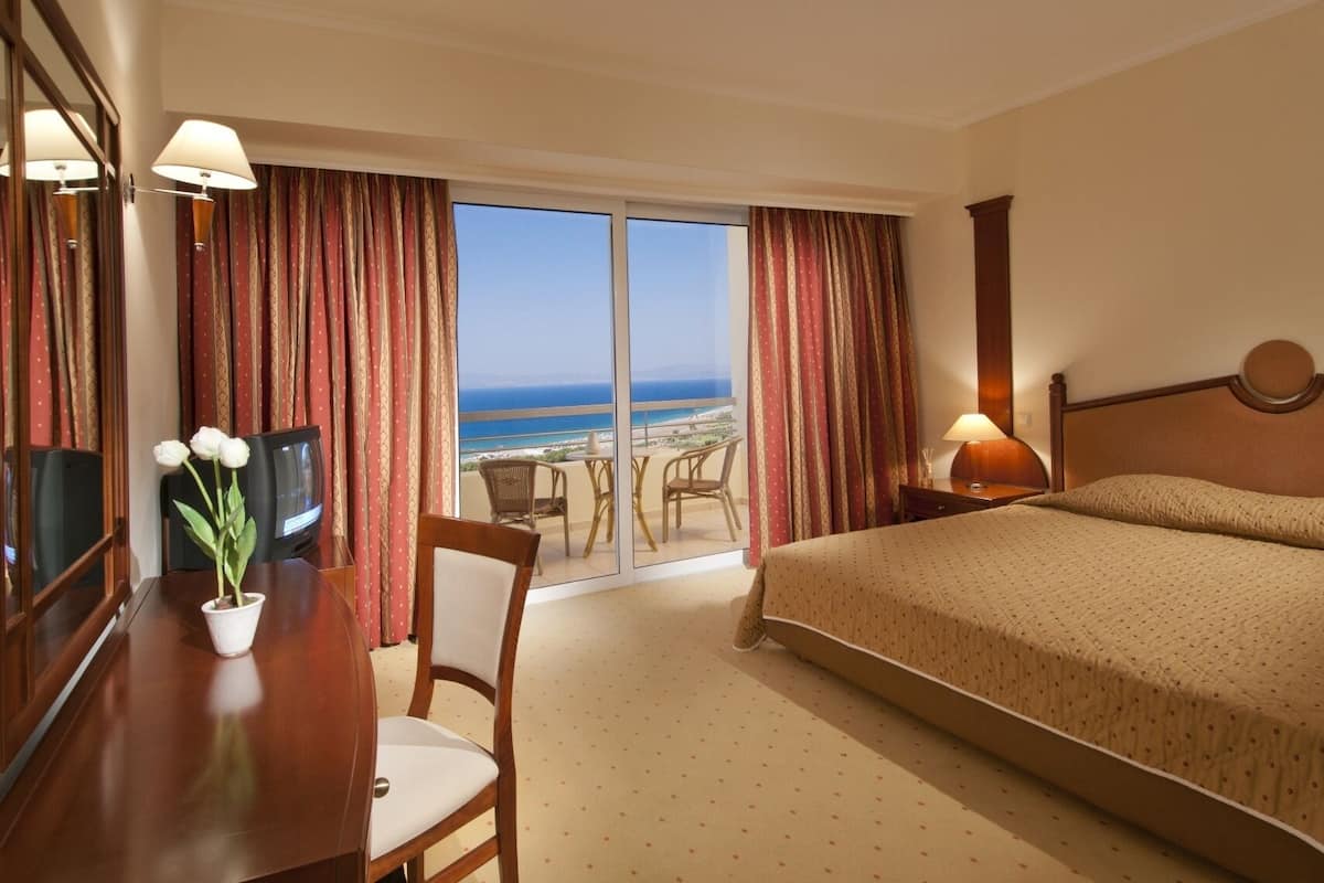 A hotel room with a bed, a desk with a lamp, a chair, a TV, and a view of the ocean.