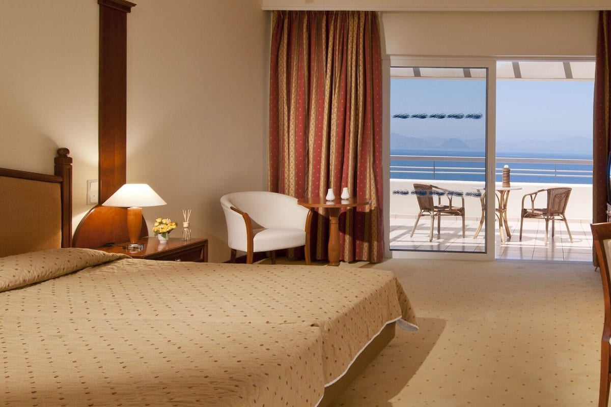 Een hotelkamer met een bed, een stoel, een tafel, een lamp en uitzicht op het strand.