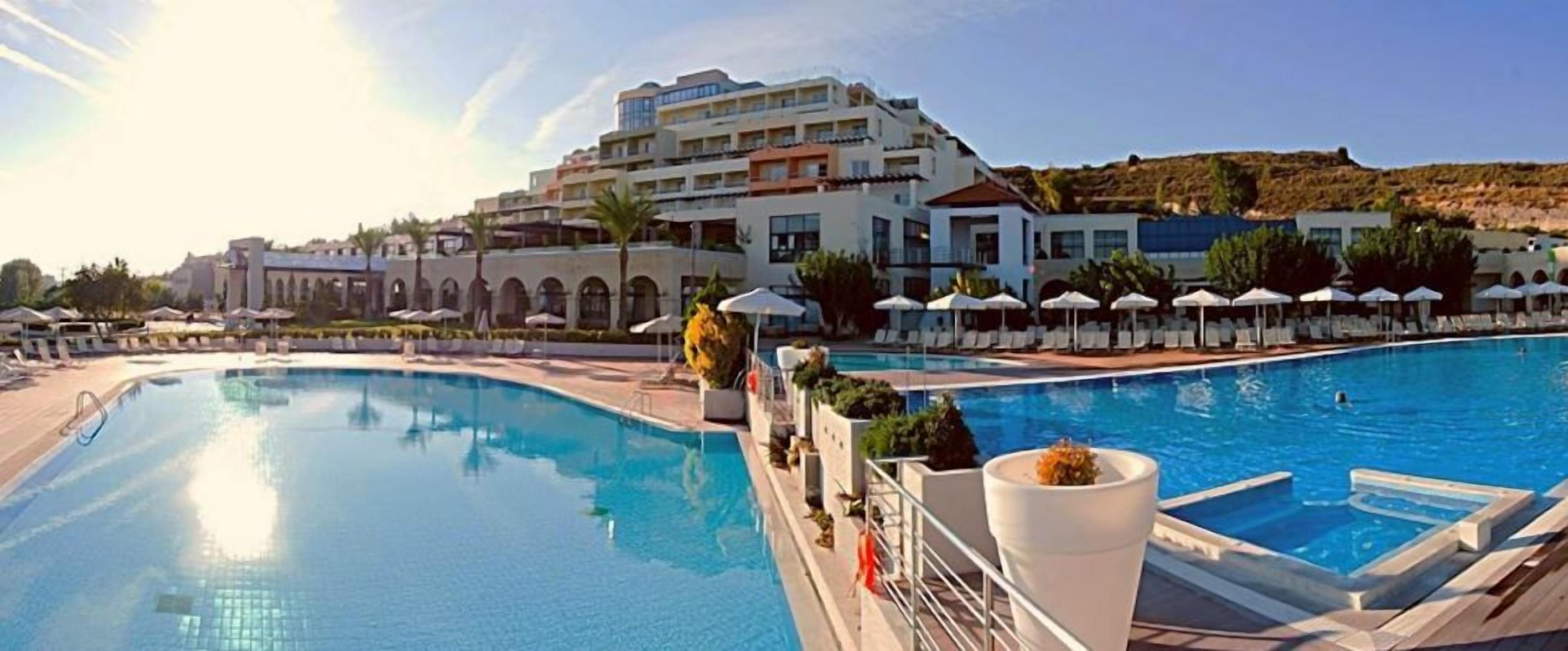 A swimming pool with lounge chairs and umbrellas, a large building with multiple balconies, and a clear sky.