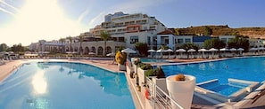 A swimming pool with lounge chairs and umbrellas, a large building with multiple balconies, and a clear sky.