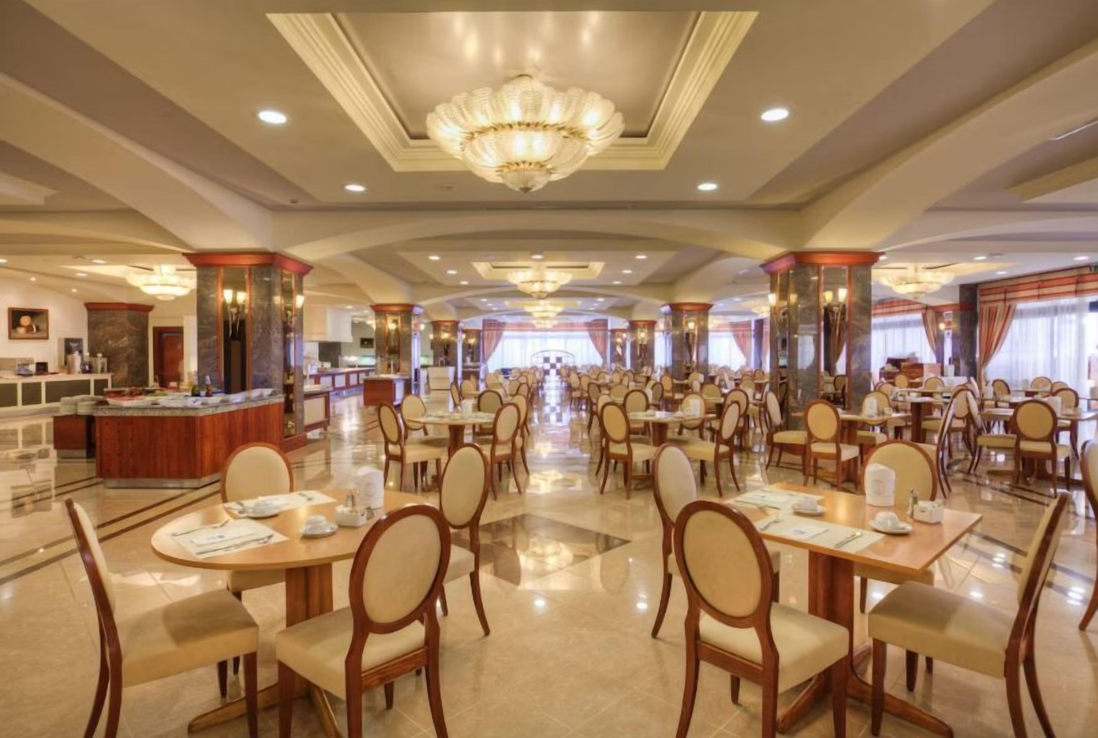 A spacious hotel dining area with multiple tables and chairs, a central bar, and a chandelier.
