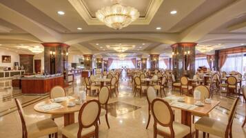A spacious hotel dining area with multiple tables and chairs, a central bar, and a chandelier.
