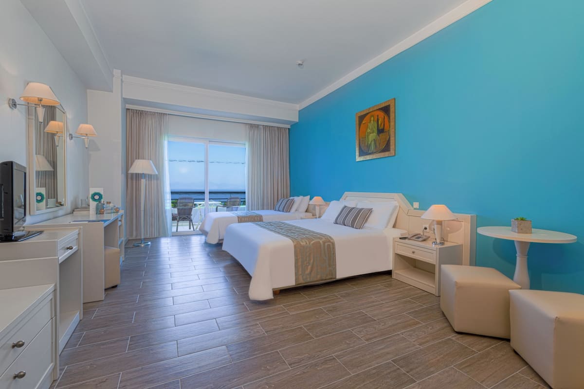 A hotel room with two beds, a desk, a chair, a TV, and a view of the sea.