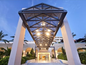 Een moderne hotelentree met een geometrisch glazen dak, een goed verlichte lobby en palmbomen op de achtergrond.