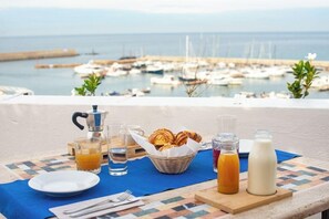 En frukostuppsättning med croissanter, apelsinjuice och mjölk, uppdukad på en terrass med utsikt över en småbåtshamn.