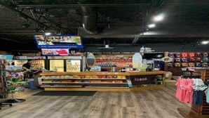 Una tienda minorista con suelo de madera, un mostrador central con televisor, estanterías con productos y una percha para ropa.
