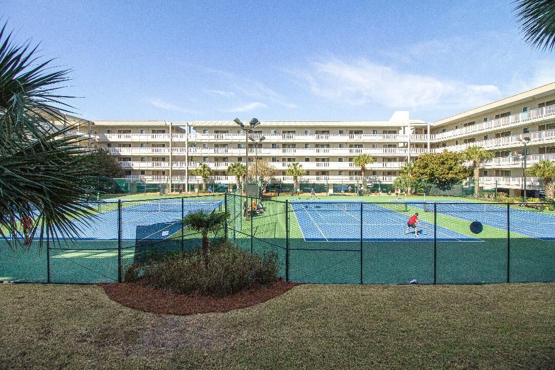 Una cancha de tenis rodeada por una valla, con un edificio de varios pisos al fondo.