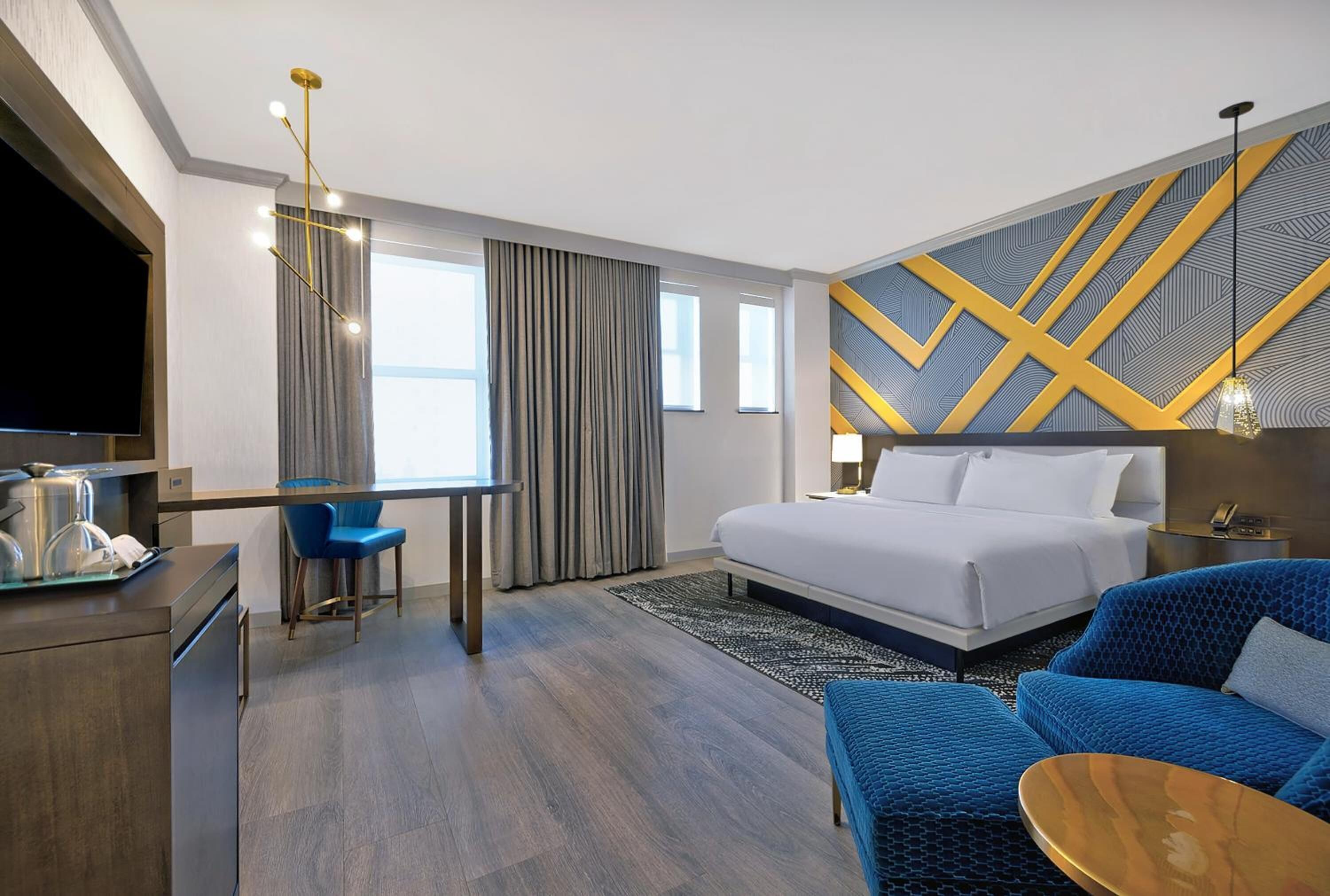 A modern hotel room with a bed, a desk with a chair, a TV, and a blue sofa.