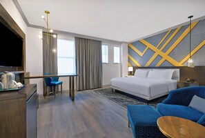 A modern hotel room with a bed, a desk with a chair, a TV, and a blue sofa. - Le Meridien St Louis Downtown (St. Louis)