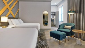 A hotel room with a large bed, a sitting area with a chair and ottoman, a small table, a mirror, and a view of the hallway and kitchen. - Le Meridien St Louis Downtown (St. Louis)