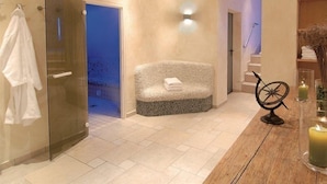 Un baño moderno con puerta de ducha de vidrio, un banco con toallas, suelo de madera y una mesita con velas.