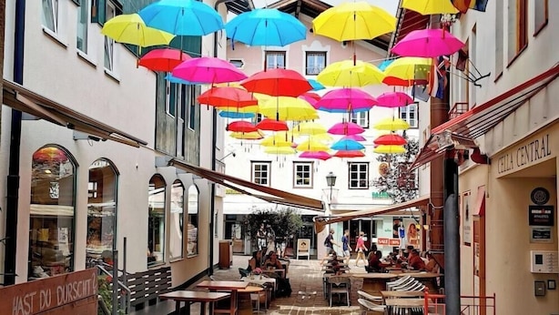 Una calle con paraguas de colores colgando sobre la cabeza, un café con terraza y un cartel con texto en alemán.
