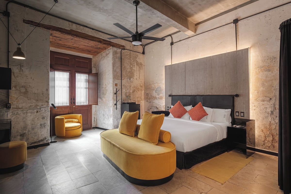 A modern bedroom with a curved sofa, a large bed with multiple pillows, a ceiling fan, and a rustic wall.