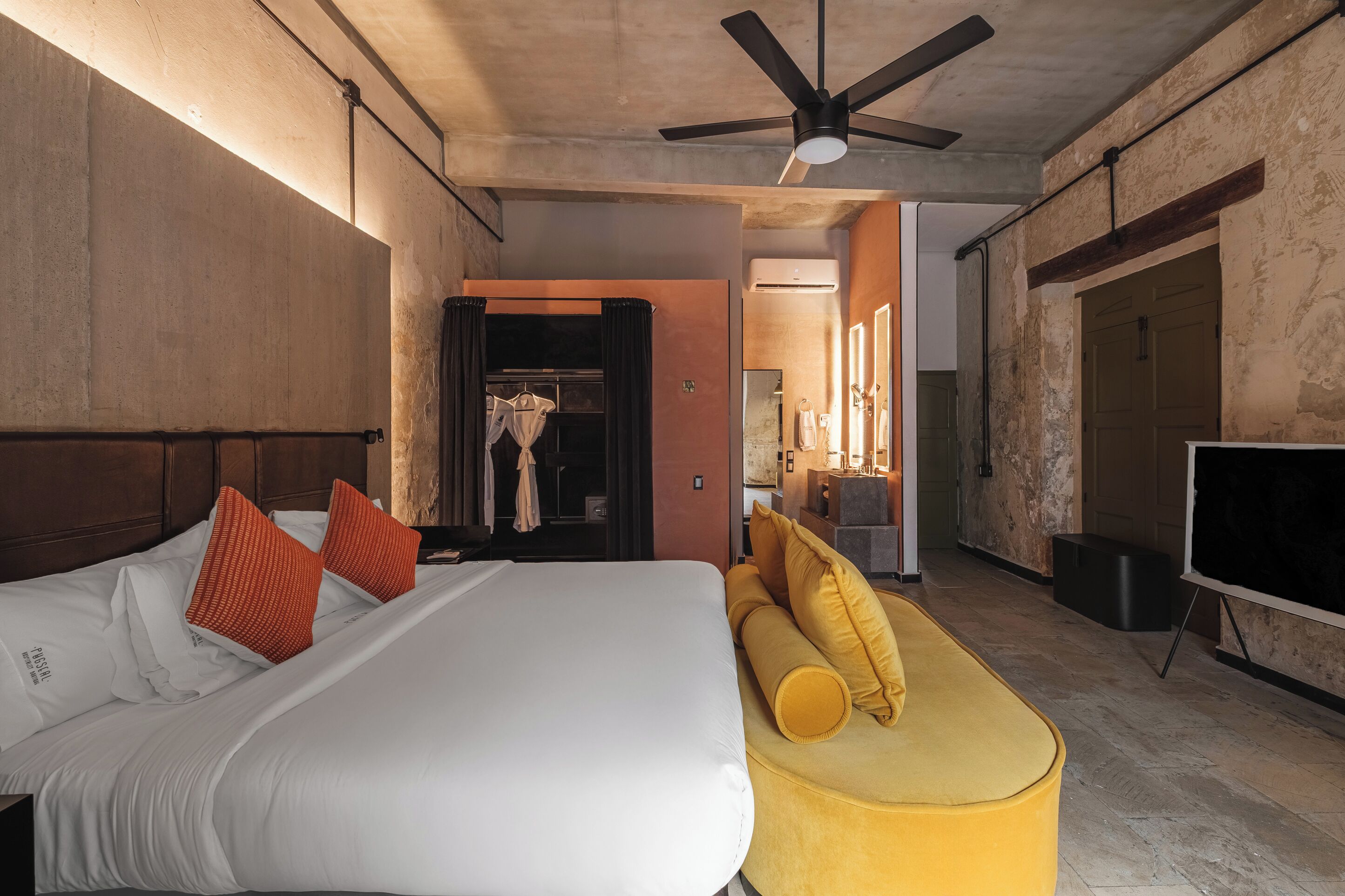 Habitació d'hotel amb un llit, un sofà groc, un ventilador de sostre i un aparell d'aire condicionat.