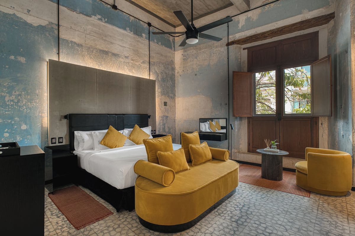 A modern hotel room with a large bed, a sofa, a small table, and a window with a view of trees.