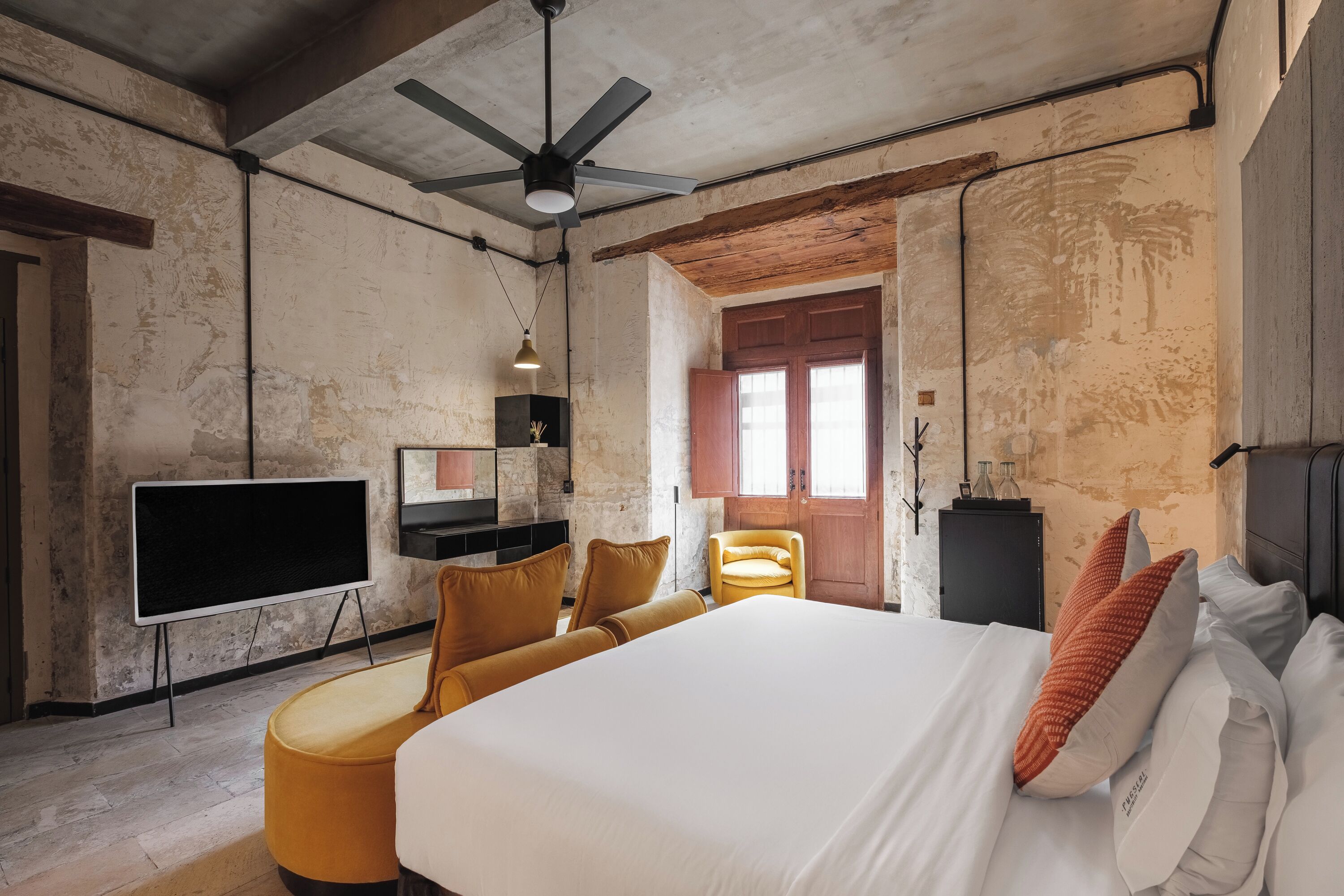 A hotel room with a large bed, a ceiling fan, a flat-screen TV, a wooden chair, and a small table.