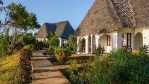 A row of thatched-roof cottages with white walls and arched doorways, surrounded by lush greenery and a clear sky. - Pearl Beach Resort by Sansi (Michamvi)