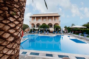 Uma piscina com espreguiçadeiras e guarda-sóis, um edifício com arcos e uma palmeira.