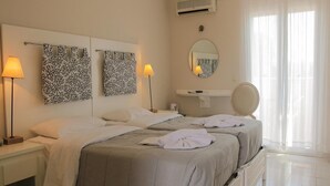 Habitación de hotel con dos camas, una mesita de noche con lámpara, un espejo y una ventana con cortinas translúcidas.