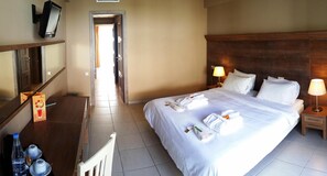 A hotel room with a large bed, two bedside tables with lamps, a desk with a flat-screen TV, a chair, and a door leading to a corridor. - Valley Village (Faistos)
