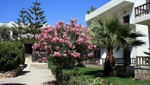 A white building with a balcony, surrounded by a garden with pink flowers and a palm tree. - Valley Village (Faistos)