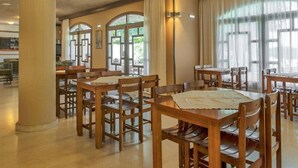 A dining area with wooden tables and chairs, large windows with curtains, and a view of the outdoors. - Valley Village (Faistos)