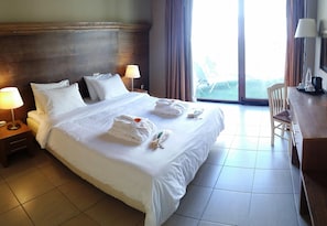 A hotel room with a large bed, two bedside lamps, a wooden headboard, a desk with a chair, and a view through the window. - Valley Village (Faistos)