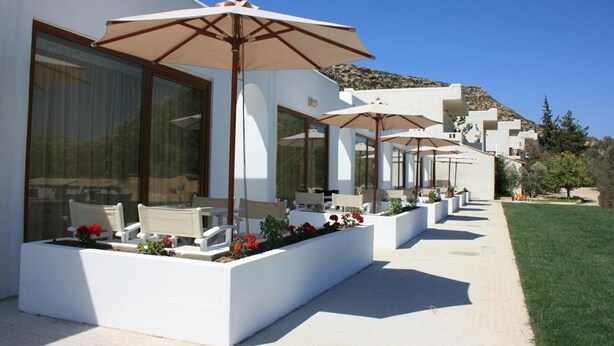A modern outdoor seating area with white planters and beige umbrellas, overlooking a landscaped garden and a hillside. - Valley Village (Faistos)