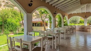 An outdoor patio with white tables and chairs, surrounded by arches and greenery. - Valley Village (Faistos)