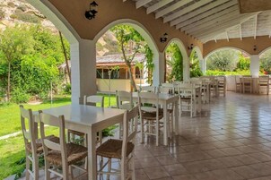 Un patio exterior con mesas y sillas blancas, rodeado de arcos y follaje.