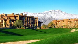 Un campo de golf con montañas al fondo y un complejo de edificios con varias plantas y balcones.