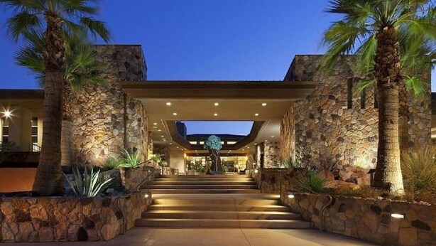Entrada de hotel con fachada de piedra, palmeras y un paseo techado que conduce al edificio principal.