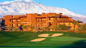 Un campo de golf con montañas al fondo y un edificio de varios pisos con balcones.