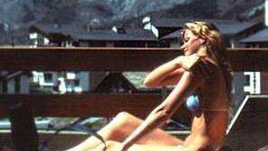 Una mujer en traje de baño sentada en una silla de terraza con una computadora portátil, con vistas a un paisaje montañoso.