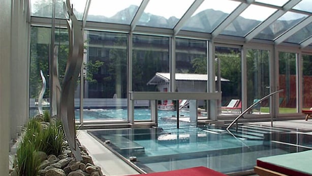 A glass-enclosed pool area with a view of mountains and a small building in the background. - Impuls Hotel Tirol (Bad Hofgastein)