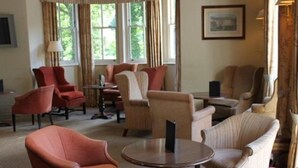 Una habitación con varias zonas de estar, incluyendo sillones y sofás, dispuestos alrededor de una mesa central. Hay ventanas amplias con cortinas, un cuadro en la pared y una lámpara sobre una mesita auxiliar.