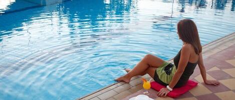 Une femme est assise au bord d’une piscine, un verre à la main et un chapeau sur la tête.