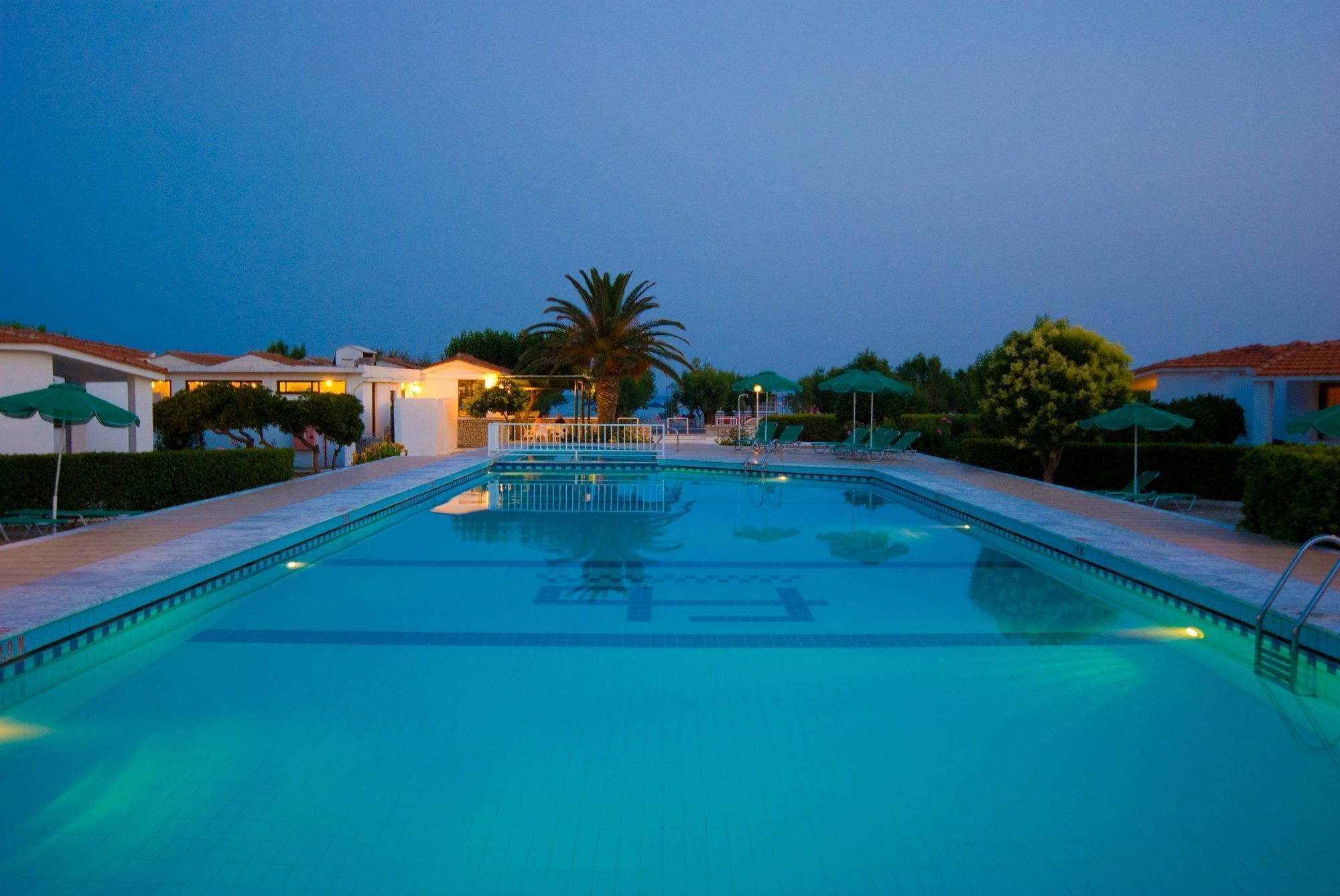 A swimming pool with a clear water surface, surrounded by lounge chairs and umbrellas, with buildings in the background.