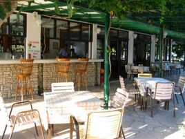 Un café al aire libre con sillas y mesas de mimbre, una zona de bar con una persona detrás del mostrador y una pared de piedra.