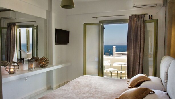 Un dormitorio con cama, televisión, balcón con vistas al mar y ventana con cortinas.