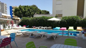 Un área de piscina con sillones de descanso y sombrillas, rodeada de edificios y follaje.