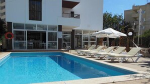 Una piscina con sillones de playa y sombrillas, rodeada por un edificio con amplios ventanales y balcones.