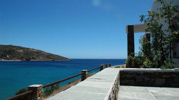 Un paseo marítimo costero con barandilla de piedra, que lleva a un edificio con fachada de piedra y vistas al mar.