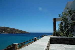 A coastal walkway with a stone balustrade, leading to a building with a stone facade and a view of the sea. - Akrotiraki (Mykonos)